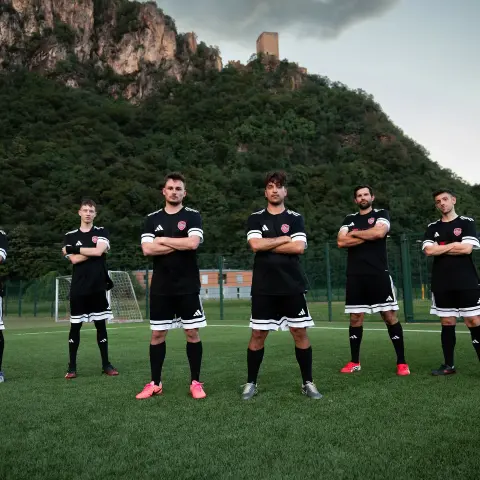 Sechs Spieler in schwarzen Trikots mit roten Wappen stehen mit verschränkten Armen auf dem Kunstrasen vor einem bewaldeten Berg mit Burgruine.