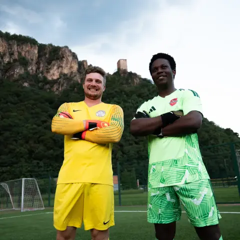 Zwei Torhüter des AFC Terlan in gelbem und grünem Trikot stehen mit verschränkten Armen lächelnd auf dem Kunstrasen vor einer Burgruine.