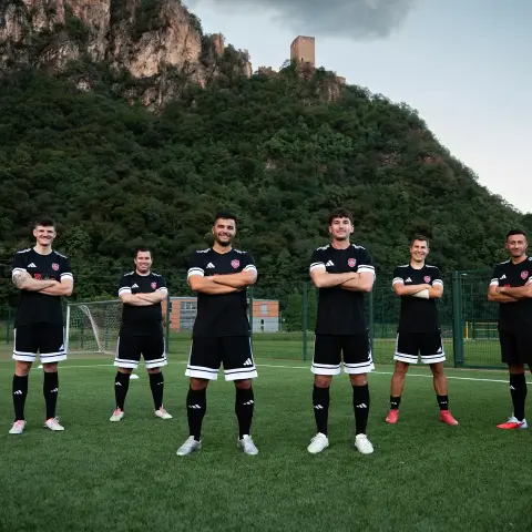 Sechs Spieler in schwarzen Trikots mit roten Wappen stehen mit verschränkten Armen auf dem Kunstrasen vor einem bewaldeten Felsen mit Burgruine.