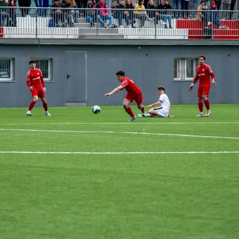 Spielszene auf dem Kunstrasen: Ein roten Spieler spielt den Ball, während ein weißer Gegenspieler am Boden rutscht, dahinter Zuschauer auf der Tribüne.