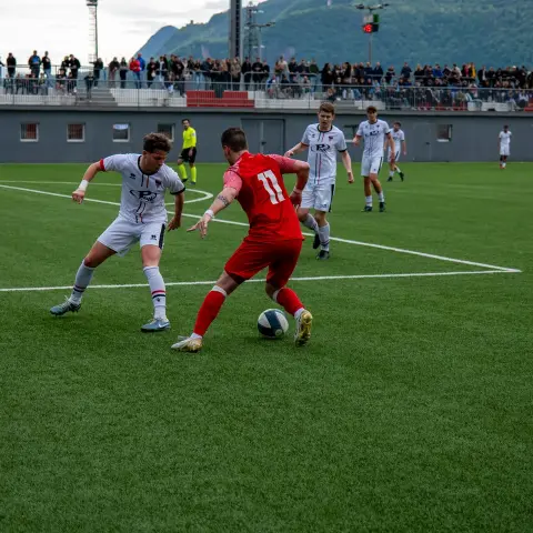 Spielszene: Ein Spieler in Rrot mit Nummer 11 führt den Ball am Gegner in Weiß vorbei, Zuschauer am Spielfeldrand im Hintergrund.