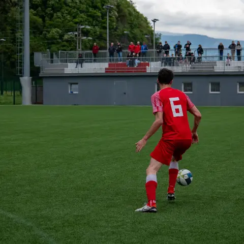 Spieler mit der Nummer 6 in roten Trikot führt den Ball auf dem Rasen, im Hintergrund Zuschauer auf der Tribüne und ein zweiter roter Spieler.