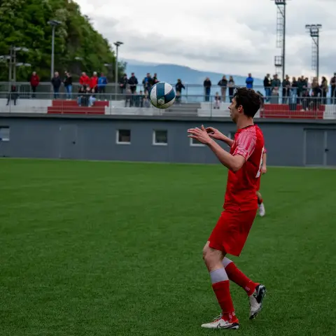 Spieler in roter Trikotkombination jongliert auf dem Kunstrasen den Ball vor einer Tribüne voller Zuschauer.