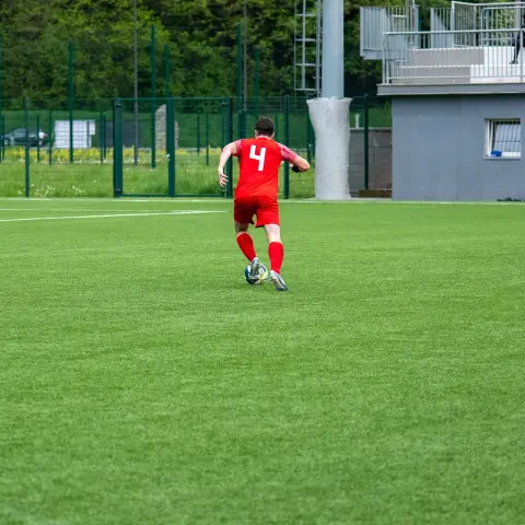 Spielszene auf dem Kunstrasen: Ein Spieler in roter Trikotnummer 4 führt den Ball, während zwei Gegenspieler in Weiß im Hintergrund und am linken Rand folgen.