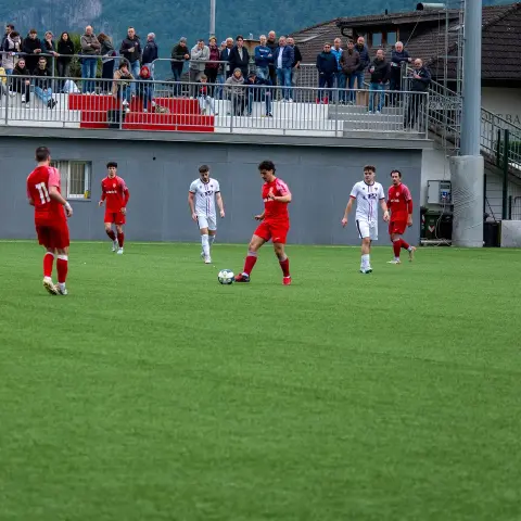 Spielszene auf dem Kunstrasen: Ein Spieler in Rot führt den Ball, Gegner in Weiß eilen herbei, im Hintergrund sitzt Publikum auf der Tribüne.