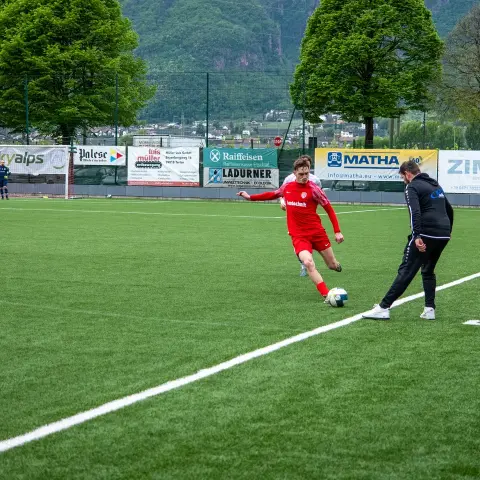 Spieler in roter Trikot mit Bautechnik-Aufdruck führt den Ball am Spielfeldrand, daneben ein Betreuer in schwarzer Jacke, im Hintergrund Werbebanden.