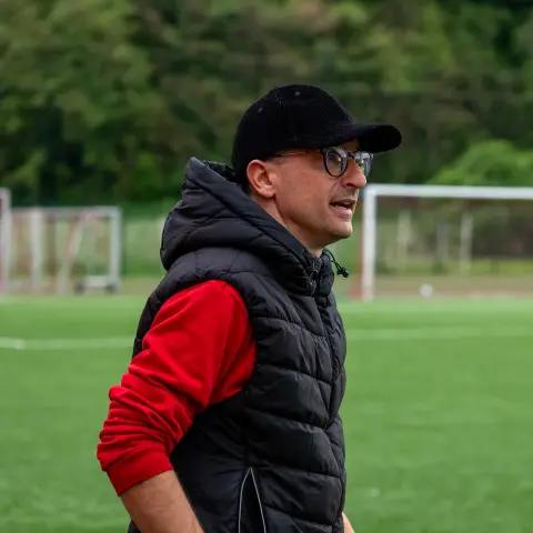 Trainer mit schwarzer Kappe, Brille und schwarzer Steppweste auf einem Kunstrasenplatz, im Hintergrund zwei Fußballtore.