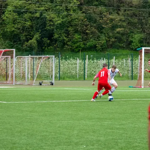 Spielszene auf dem Kunstrasen: Ein Spieler in Rot mit der Nummer 11 deckt den Ball gegen einen Angreifer in Weiß, im Hintergrund zwei Tore.