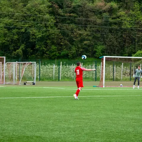 Spieler in roten Trikot mit Nummer 11 schießt den Ball auf dem Kunstrasenplatz, im Hintergrund steht ein Torwart im grünen Trikot vor dem Tor.