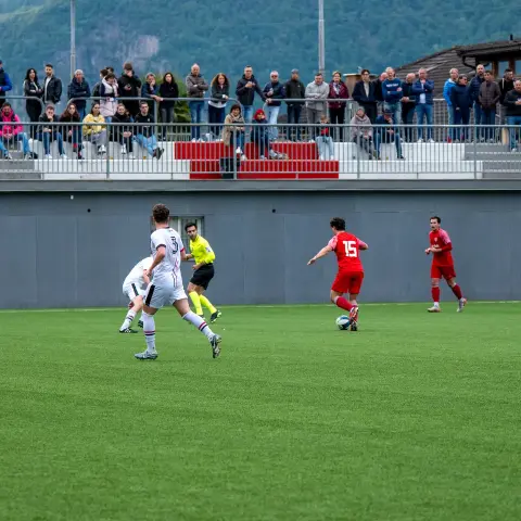 Spielszene auf dem Kunstrasen: Ein Spieler in Rot mit Ball wird von Gegenspielern in Weiß begleitet, im Hintergrund schaut Publikum zu.