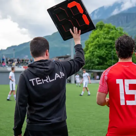 Auswechselung beim AFC Terlan: Ein Mann im schwarzen Hoodie mit Terlan-Schriftzug hält die elektronische Wechseltafel hoch, daneben steht Spieler Nummer 15 in Rot.