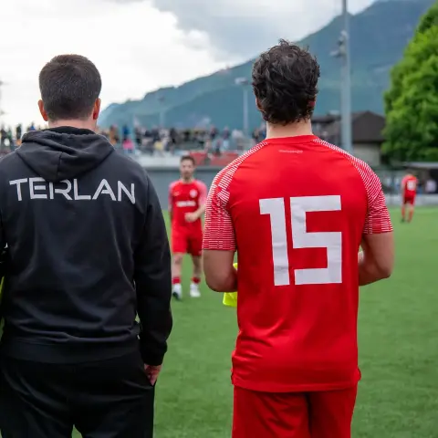 Ein Trainer im schwarzen Hoodie mit Aufschrift TERLAN und ein Spieler in roter Trikot-Nummer 15 blicken gemeinsam auf das Spielfeld.