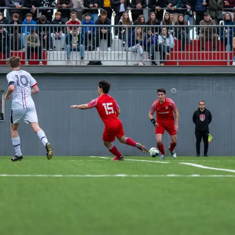 Rot gekleideter Spieler Nr. 15 führt den Ball am Spielfeldrand, ein Gegenspieler in Weiß mit Nr. 10 läuft im Hintergrund mit, Zuschauer auf der Tribüne.