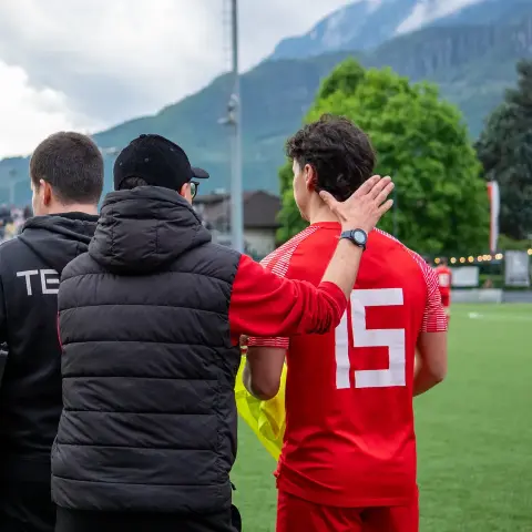 Ein Spieler in roter Trikotnummer 15 wird von zwei Betreuern in dunklen Jacken über den Platz begleitet, im Hintergrund Zuschauer und Berge.