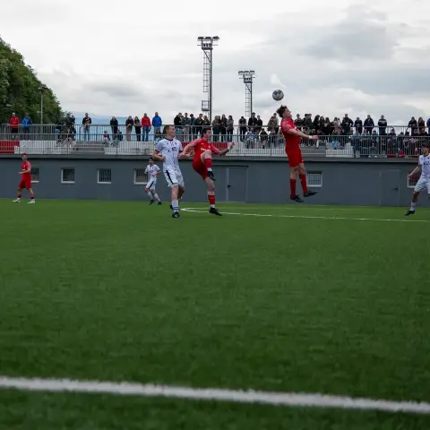 Spielszene auf dem Kunstrasenplatz: Ein roter Spieler springt am Strafraum zum Kopfball, mehrere weiß gekleidete Gegenspieler stehen in der Nähe, Zuschauer am Zaun im Hintergrund.