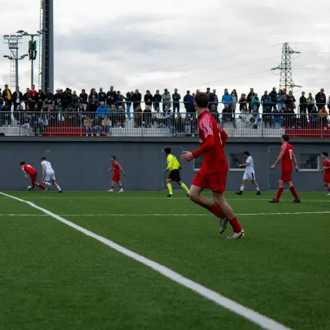 Spielszene auf einem Kunstrasenplatz: Rot gekleidete Spieler des AFC Terlan laufen einem Ball nach, im Hintergrund sitzt Publikum auf der Tribüne.