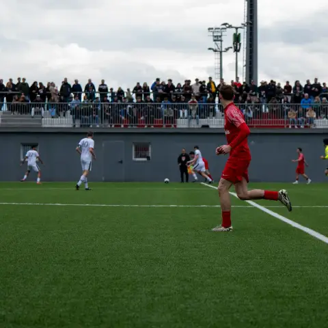 Spieler in roter Trikot läuft über den Rasen während eines Spiels, im Hintergrund sitzen Zuschauer auf der Tribüne.