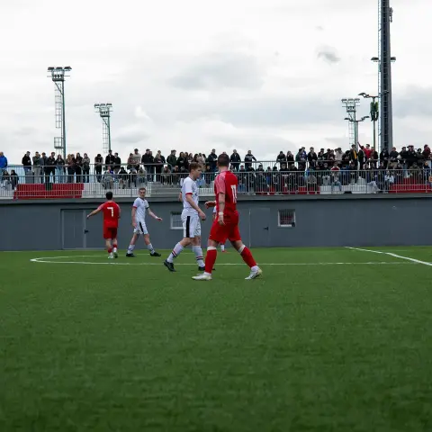 Spielszene auf dem Kunstrasen: Spieler in roten Trikots und ein Spieler in Weiß, im Hintergrund Zuschauer auf der Tribüne.
