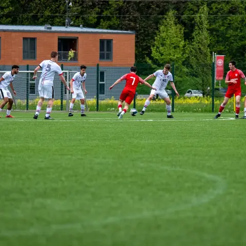 Spielszene auf dem Kunstrasen: Ein Spieler in Rot mit Nummer 7 führt den Ball gegen zwei Gegner in Weiß.
