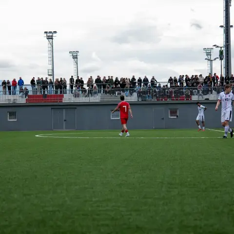 Fußballspiel auf einem grünen Kunstrasenplatz: Ein Spieler in Rot wartet, während zwei Spieler in Weiß zurücklaufen, im Hintergrund Zuschauer auf der Tribüne.
