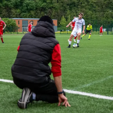 Spielszene: Ein Spieler in Weiß führt den Ball, während ein Trainer in Schwarz am Spielfeldrand kniet und das Spiel verfolgt.