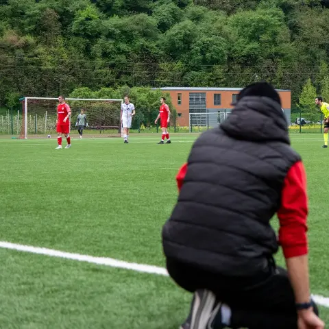 Spielszene auf dem Kunstrasen: Ein Zuschauer in dunkler Steppweste kniet am Spielfeldrand, im Hintergrund spielen Mannschaften in Rot und Weiß, der Schiedsrichter in Gelb.