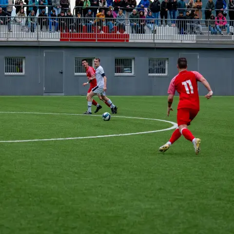 Spielszene auf einem Kunstrasenplatz: Ein Spieler in Rot mit der Nummer 11 läuft dem Ball hinterher, zwei Gegner in Weiß und Rot folgen, Zuschauer auf der Tribüne.