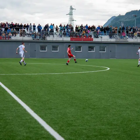 Spielszene auf dem Kunstrasenplatz: Ein roter Spieler führt den Ball, zwei weiße Gegenspieler laufen mit, im Hintergrund stehen zahlreiche Zuschauer auf der Tribüne.
