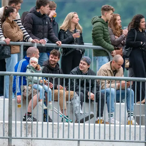 Zuschauer stehen und sitzen hinter einem Metallgeländer am Spielfeldrand, viele mit Getränken in der Hand, darunter eine Familie mit Kleinkind.