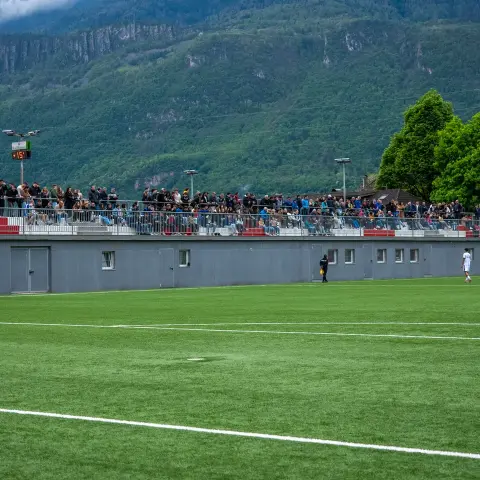 Gut besuchte Tribüne mit roten und weißen Sitzreihen vor grauem Stützmauerbau, dahinter Berghänge; auf dem Kunstrasen stehen nur wenige Spieler.