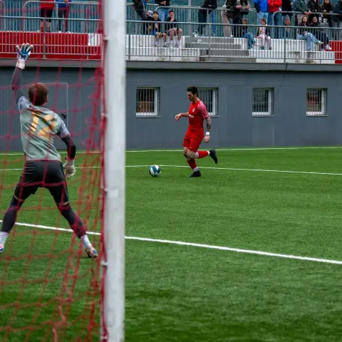 Spielszene: Ein Spieler in Rot führt den Ball Richtung Tor, der Torhüter in Grau wartet links im Kasten.