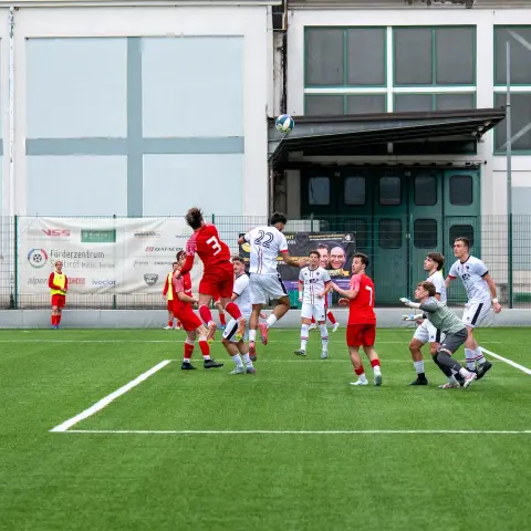 Spielszene: Weißer Spieler (22) steigt zum Kopfball hoch, davor Gegenspieler in Rot und der Torwart in Grau im Strafraum.