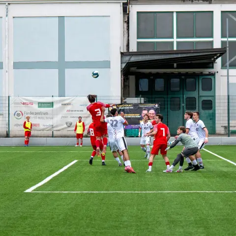Spielszene: Mehrere Spieler in Rot und Weiß kämpfen im Strafraum um einen Kopfball, daneben ein Torwart in Grau auf dem Kunstrasen.