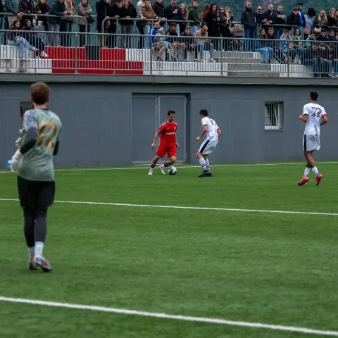 Spielszene: Roter Spieler führt den Ball gegen zwei Gegenspieler in Weiß, im Hintergrund Zuschauer auf der Tribüne.