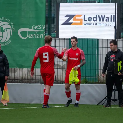 Zwei Spieler des AFC Terlan in roten Trikots klatschen sich am Spielfeldrand ab, im Hintergrund ein Banner und eine LED-Wand.