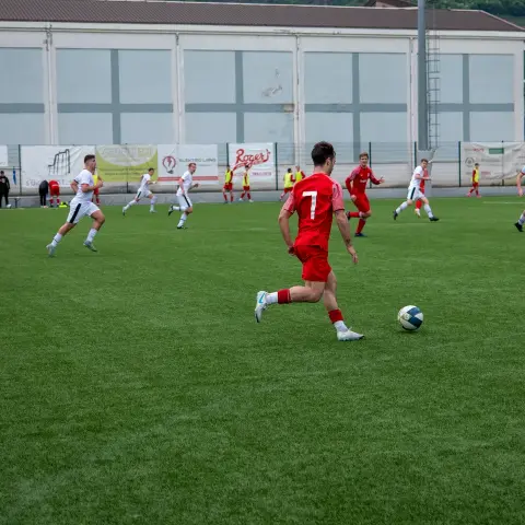Spieler in rotem Trikot mit der Nummer 7 führt den Ball auf dem Kunstrasen, Gegenspieler in Weiß laufen mit; im Hintergrund Banden mit Sponsorenwerbung.