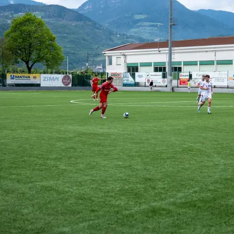 Spielszene auf dem Kunstrasen: Ein Spieler in Rot führt den Ball, Gegenspieler in Weiß, Werbebanden am Spielfeldrand.