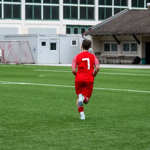 Spieler in roter Trikot mit Nummer 7 läuft auf dem Kunstrasenplatz, von hinten fotografiert; im Hintergrund Tornetze und ein Vereinsgebäude.