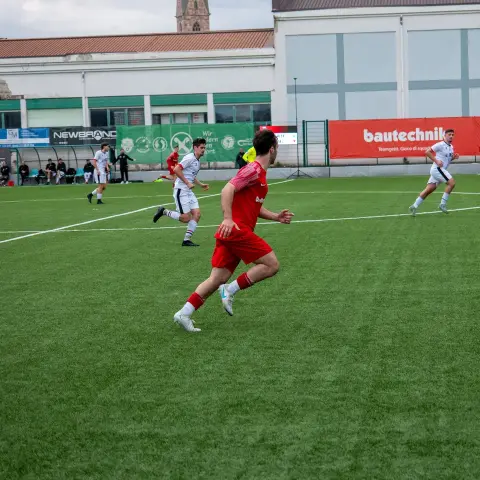 Spieler in roter Trikot und roter Hose läuft auf dem Kunstrasen einem Ball nach, im Hintergrund Spieler in Weiß und Werbebanden, darunter bautechnik.