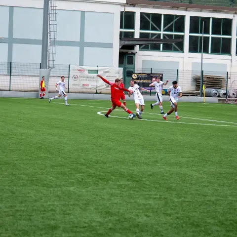 Spielszene auf dem Kunstrasen: Ein Spieler in Rot kämpft mit dem Ball gegen zwei Gegner in Weiß, im Hintergrund ein Banner mit Sponsoren.