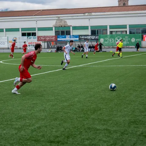 Spielszene: Ein Spieler in roter Trikot läuft mit dem Ball über den Rasen, während ein Gegenspieler in Weiß folgt.