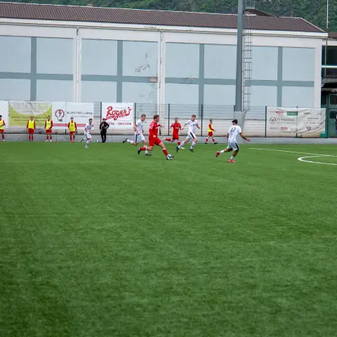 Spielszene auf dem Kunstrasen: Ein Spieler in Rot führt den Ball, zwei Gegner in Weiß folgen, im Hintergrund Werbebanden.