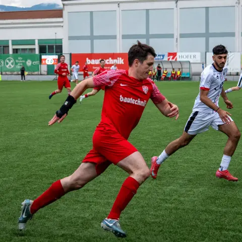 Spielszene: Ein Spieler in roter Trikot mit Bautechnik-Sponsor läuft mit dem Ball am Fuß an einem Gegner in Weiß vorbei auf dem Kunstrasenplatz.