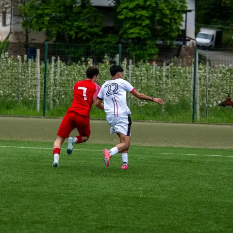 Zwei Fußballspieler im Zweikampf auf dem grünen Kunstrasen, links in Rot mit Nummer 7, rechts in Weiß mit Nummer 22, der Ball rollt rechts am Spielfeldrand.