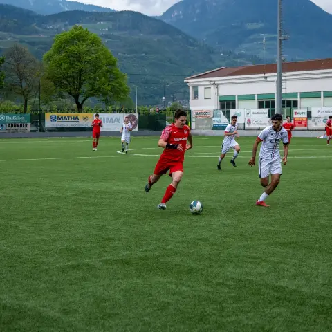 Spielszene auf dem Kunstrasen: Ein Spieler in roter Trikot führt den Ball, ein Gegner in Weiß sowie Banden mit Sponsorenwerbung im Hintergrund.