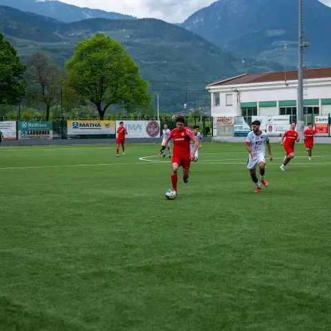 Spielszene auf einem Kunstrasenplatz: Ein Spieler in roter Trikot führt den Ball, ein Gegner in Weiß verfolgt ihn, im Hintergrund Banden mit Sponsorenlogos.