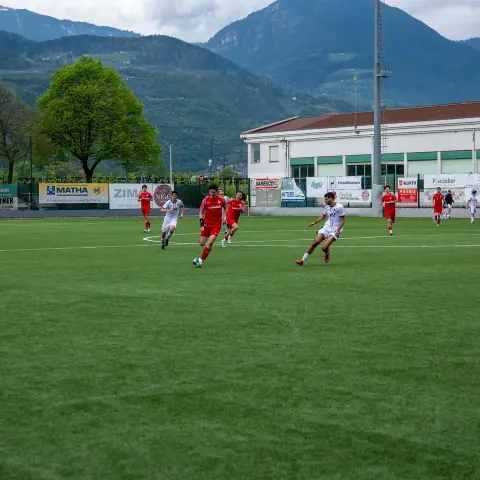 Spielszene auf dem Kunstrasenplatz: Rote Mannschaft und Gegner in Weiß laufen dem Ball nach, im Hintergrund Bergkulisse und Bandenwerbung.
