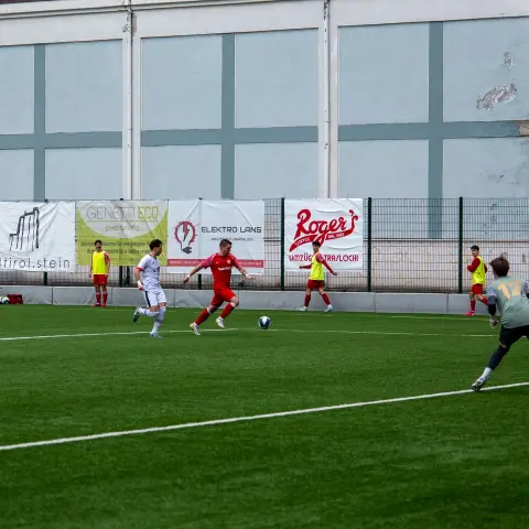 Spielszene auf dem Kunstrasen: Roter Spieler führt den Ball gegen einen Gegner in Weiß, im Hintergrund Sponsorenbanden.