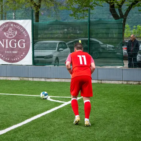 Spieler in roten Trikot mit Nummer 11 steht hinter dem Ball an der Eckfahne auf dem Kunstrasen, im Hintergrund Banden mit Werbung der Metzgerei Nigg.