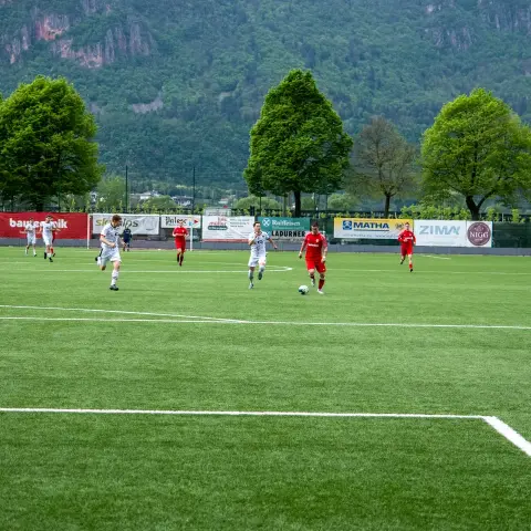 Spielszene auf dem Kunstrasen: Rote Spieler in Trikots und weiße Gegenspieler laufen über das Feld, im Hintergrund Werbebanden und bewaldete Berge.